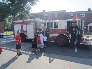 Children at Sullivan Apartments line up to see the interior of a fire truck. Captain Robert Fancy stands at the passenger door.